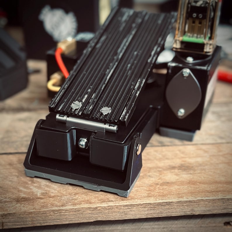 May include: A black metal foot pedal with a ribbed surface and a hinge. The pedal has a gray rubber base and is designed for use with a machine.