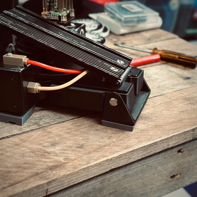 May include: A black foot pedal with a textured surface and a red and yellow cable attached. The pedal is on a wooden surface.