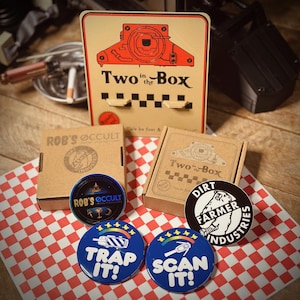 Puede incluir: Una colección de cuatro pines redondos con varios diseños. Los pines presentan el texto "Trap It!" y "Scan It!" junto con un gráfico de un coche y un escáner. Los pines se muestran sobre una superficie a cuadros rojos y blancos con dos cajas marrones que muestran el texto "Rob's Occult" y "Two in the Box".