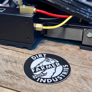 May include: A black and white sticker with the text "Dirt Farmer Industries" and a cartoon image of a bear wearing a hat. The sticker is on a wooden surface.