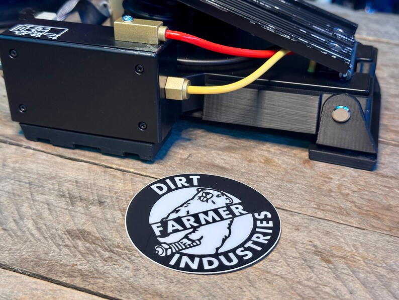 May include: A black and white sticker with the text "Dirt Farmer Industries" and a cartoon image of a bear wearing a hat. The sticker is on a wooden surface.