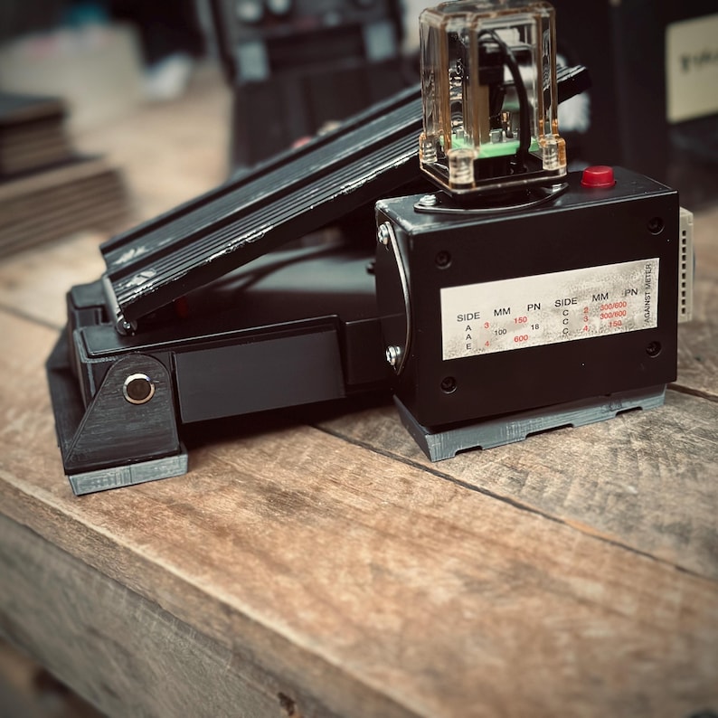 May include: A black foot pedal with a gray base and a clear plastic box on top. The box has a red button and a label that reads "SIDE MM PN SIDE MM PN 150 A 100 18 C C 300100 600 AGAINST METER".