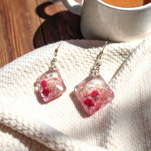 May include: Dangle earrings with clear, diamond-shaped resin pendants containing dried pink flowers. The earrings have gold-coloured hooks and crystal accents. They sit on a cream knitted fabric. A mug of tea is in the background.