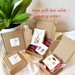 May include: Open brown gift boxes displaying jewellery, including a gold necklace with a rectangular pendant and earrings. The boxes have white labels with floral designs. Text reads "Free gift box with every order!"