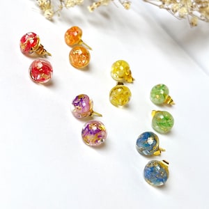May include: A selection of colourful stud earrings. Each earring has a clear, spherical resin design with dried flowers and gold flakes. The earrings have gold-coloured posts and are arranged on a white surface.