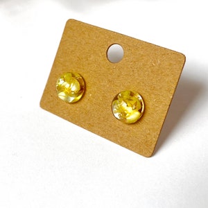 May include: A pair of round stud earrings with clear, yellow resin. The earrings contain small, dried yellow flowers and gold flakes. The earrings are displayed on a brown earring card with a hole at the top.