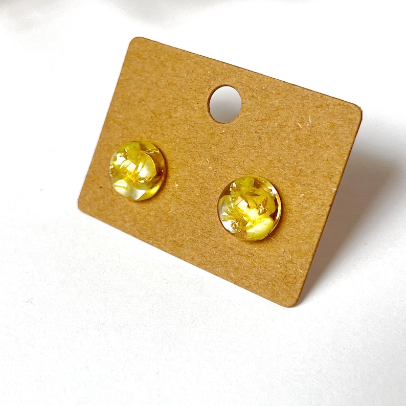 May include: A pair of round stud earrings with clear, yellow resin. The earrings contain small, dried yellow flowers and gold flakes. The earrings are displayed on a brown earring card with a hole at the top.