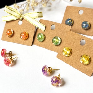 May include: A selection of colourful resin stud earrings with gold-coloured posts. The earrings feature dried flowers and gold leaf encased in clear resin. The earrings are displayed on small, brown earring cards.