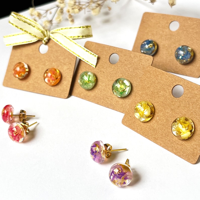 May include: A selection of colourful resin stud earrings with gold-coloured posts. The earrings feature dried flowers and gold leaf encased in clear resin. The earrings are displayed on small, brown earring cards.