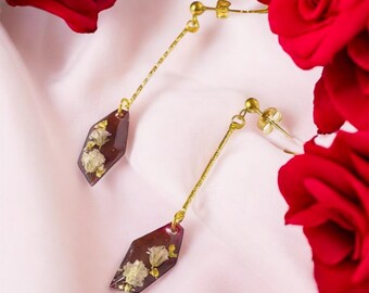 Puede incluir: Un par de pendientes colgantes con postes y cadenas doradas. Los pendientes presentan un colgante geométrico de color rojo oscuro con pequeñas flores blancas y copos de oro encapsulados en resina. El fondo incluye rosas rojas y tela rosa.