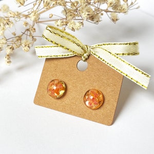 May include: Round stud earrings featuring a floral design set in clear resin. Presented on a brown earring card, they are decorated with a cream ribbon with gold edging. The background is white.