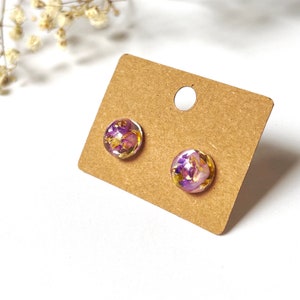 May include: A pair of round stud earrings with a floral design. The earrings feature a clear resin dome encasing purple and gold-coloured dried flowers. They are displayed on a brown earring card against a white background.