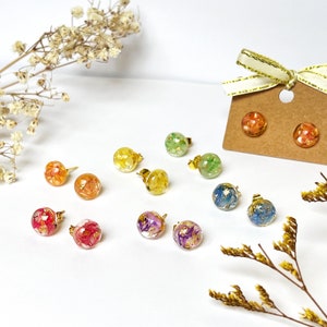 May include: A selection of stud earrings with gold posts. Each earring has a clear, spherical resin design, incorporating colourful dried flowers and gold leaf. The earrings are presented on a white surface, with dried floral decorations.