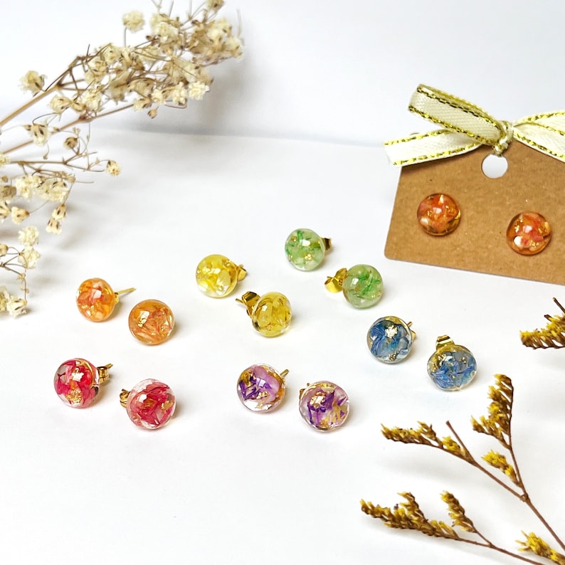May include: A selection of stud earrings with gold posts. Each earring has a clear, spherical resin design, incorporating colourful dried flowers and gold leaf. The earrings are presented on a white surface, with dried floral decorations.