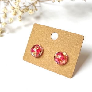 May include: A pair of clear, round stud earrings with pink floral inclusions and gold flakes, presented on a brown earring card. The earrings are set against a white background.