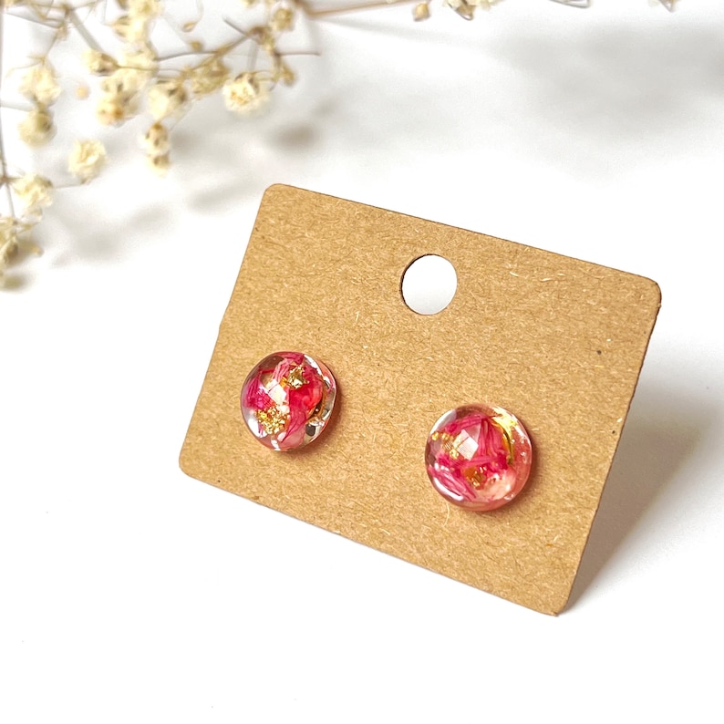 May include: A pair of clear, round stud earrings with pink floral inclusions and gold flakes, presented on a brown earring card. The earrings are set against a white background.