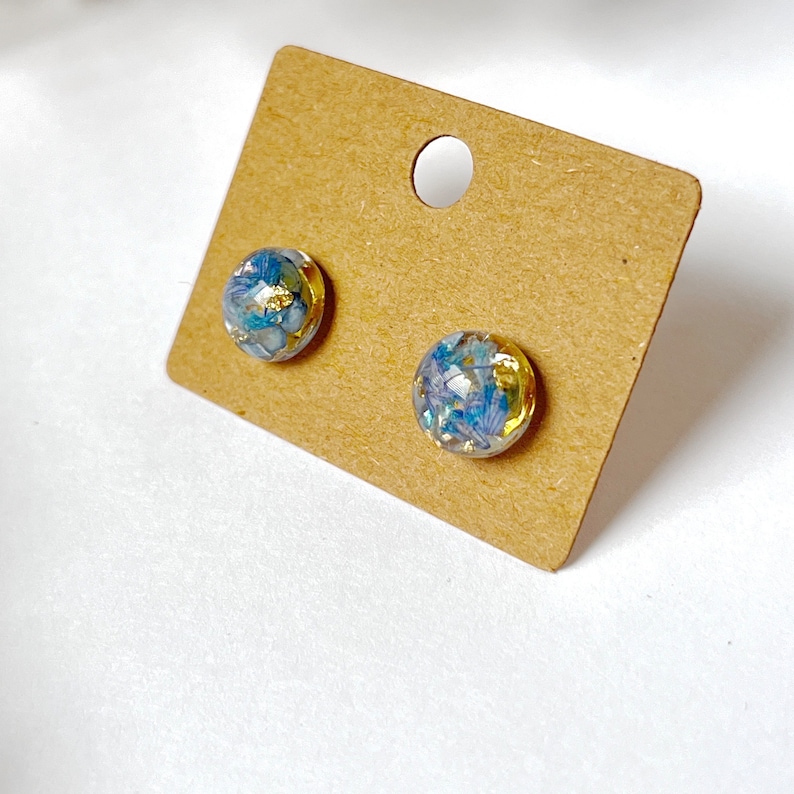 May include: A pair of round stud earrings. Each earring has a clear resin dome with blue and gold floral inclusions. The earrings are displayed on a brown cardboard earring card with a circular hole.