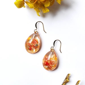 May include: A pair of teardrop earrings with gold-coloured hooks. The clear resin earrings contain dried orange and yellow flowers and gold flakes. The earrings are displayed on a white surface with yellow flowers in the background.