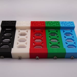 Könnte beinhalten: Fünf 3D-gedruckte Kunststoffständer in Schwarz, Weiß, Rot, Grün und Blau. Jeder Ständer hat ein Gittermuster und den Text "ZLR TECH" auf der Oberseite.