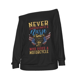 May include: Black off-the-shoulder long-sleeve shirt. The shirt features the text "NEVER UNDERESTIMATE A Nurse WHO RIDES A MOTORCYCLE" in gold, blue, and red. A motorcycle engine graphic with wings is centered on the front.