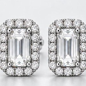 May include: A pair of rectangular stud earrings featuring a large emerald-cut clear center stone surrounded by a halo of smaller round brilliant-cut clear stones set in a polished silver-toned metal frame with a classic prong setting design.