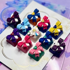 May include: A collection of eleven square mechanical keyboard keycaps, each featuring a sculpted white heart base topped with a colourful ribbon bow. The bows come in various shades including purple, blue, red, yellow, pink, and green.