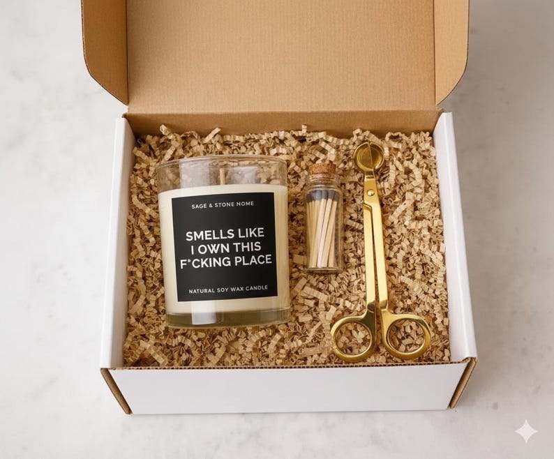 May include: A gift box containing a glass candle with the text "Smells Like I Own This F*cking Place", a small glass jar of matches, and gold-colored wick trimmers. The candle is a natural soy wax candle.