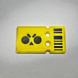 May include: A yellow plastic key card with a black and white graphic of a face with two eyes and a triangle nose. The card has several holes and a black and white barcode on the right side.