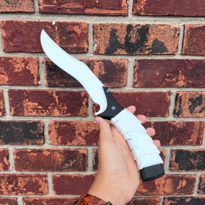 May include: A white and black plastic knife with a curved blade and a textured handle. The knife is held in a person's hand.