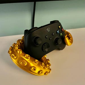 May include: A black video game controller is resting in a gold 3D printed octopus stand. The controller has buttons and joysticks. The stand is shaped like an octopus with tentacles.
