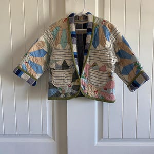 May include: A quilted jacket constructed from various fabric patches in shades of blue, tan, and white. The garment features a shawl collar, two front patch pockets with horizontal stripes, and green trim along the edges and sleeve cuffs.