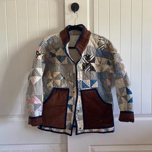 May include: A quilted jacket constructed from various patchwork fabric squares in shades of blue, white, and tan. The garment features a brown collar, brown patch pockets, and dark blue trim along the edges, cuffs, and front button closure.