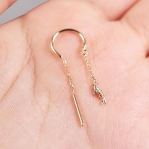 May include: A gold-toned threader earring featuring a delicate chain design. One end of the chain terminates in a slender, polished metal bar, while the other end features a small, detailed charm shaped like a swimming koi fish.