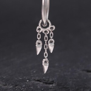 Peut inclure: Une seule boucle d'oreille argentée avec un design de chaîne pendante et de cerceau. La boucle d'oreille comporte trois petits breloques pointues suspendues à de fines chaînes. La boucle d'oreille est présentée sur un fond sombre.