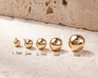 Può includere: Un set di cinque orecchini a perno con sfera dorata in diverse dimensioni, che vanno da 1,5 mm a 4 mm. Gli orecchini sono esposti su una superficie testurizzata beige chiaro.