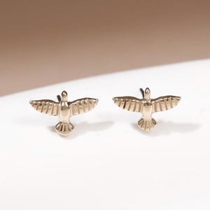 Pode incluir: Um par de pequenos brincos de pino em metal dourado com formato de pássaros em voo. Cada brinco apresenta asas estendidas com detalhes de linhas gravadas e uma cauda em leque, projetados para serem usados como um conjunto correspondente.