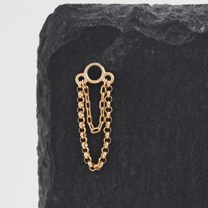 Puede incluir: Un pendiente de color dorado con un diseño de cadena. El pendiente presenta una pequeña parte superior circular con dos cadenas colgando. El pendiente se muestra sobre una superficie negra y texturizada.