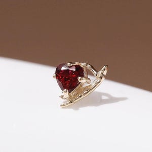 May include: A gold-toned stud earring featuring a deep red, heart-shaped gemstone held by a delicate, textured metal setting that curves around the base of the stone, creating a small, intricate piece of jewelry for the ear.