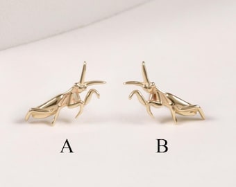 May include: Two gold-toned metal stud earrings shaped like praying mantises, labeled with the letters A and B underneath each piece. The earrings feature detailed insect anatomy with slender legs and antennae, presented as a pair of individual jewelry items.