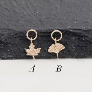 May include: Two gold-colored charms, one shaped like a maple leaf and the other like a ginkgo leaf, each with a small loop at the top. The charms are labeled "A" and "B" and are displayed against a dark, textured background.