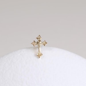 May include: A small, gold-colored cross with a detailed design. The cross features a central gem and decorative elements on each arm. It is set against a soft, white background, highlighting its intricate craftsmanship and religious symbolism.