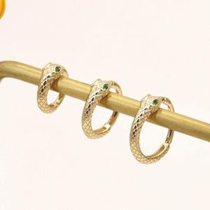 Könnte beinhalten: Drei goldene Creolen in Schlangenform mit texturierten Schuppen und kleinen grünen Augen. Die Ohrringe sind auf einem goldfarbenen Metallständer präsentiert. Ein gelbes quadratisches Abzeichen enthält den Text 14K SOLID GOLD.