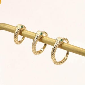 Puede incluir: Tres pendientes de aro de oro con forma de serpiente, con escamas texturizadas y pequeños ojos de piedra preciosa verde. Los pendientes se muestran en un soporte de metal dorado. Una insignia cuadrada amarilla en la esquina superior izquierda contiene el texto 14K SOLID GOLD.
