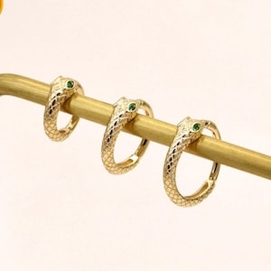 Puede incluir: Tres pendientes de aro dorados con forma de serpiente y ojos de piedra verde se exhiben en un soporte dorado. Los pendientes tienen un patrón texturizado. Un cuadrado dorado con el texto "14K SOLID GOLD" es visible.