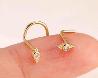 Peut inclure: Deux bijoux de nez en or ornés d'une petite pierre précieuse ronde et transparente sertie dans une base métallique avec des détails perlés. L'un possède une tige incurvée et l'autre une tige en forme de L. Les bijoux sont dorés.
