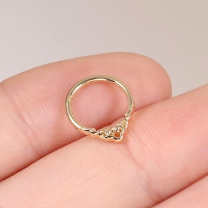 Puede incluir: Un anillo circular de tono dorado que presenta un adorno decorativo en forma de corona en el borde inferior. La pieza de joyería está diseñada con un segmento articulado para abrir y cerrar, mostrando un acabado metálico pulido.