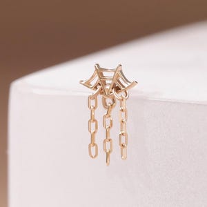 May include: A gold-toned metal earring stud featuring a geometric, fan-like top structure with three delicate, dangling chain segments suspended from the base. The piece has a polished finish and a symmetrical design with multiple small rectangular links.