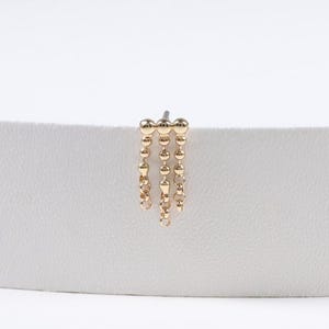 Op de afbeelding: Een goudkleurige oorbel met een pin en drie hangende strengen van kleine, ronde kralen. De oorbel is geplaatst tegen een witte achtergrond.