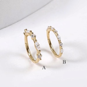 May include: Two gold-colored hoop earrings. One earring features a row of small, clear gemstones and a larger, marquise-cut stone. The other earring has a similar design, with a row of small, clear gemstones and a marquise-cut stone. The earrings are labeled A and B.