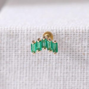 Puede incluir: Un pendiente dorado con cinco piedras rectangulares de color verde esmeralda dispuestas en forma de arco. El pendiente tiene un pequeño poste redondo. El diseño es sencillo y elegante, adecuado para diversas ocasiones.